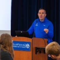 A speaker wearing a blue zip-up stands at a podium